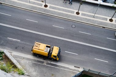 Sarı bir çöp kamyonu arka planda yayaların dolaştığı bir sokak boyunca atık topluyor..