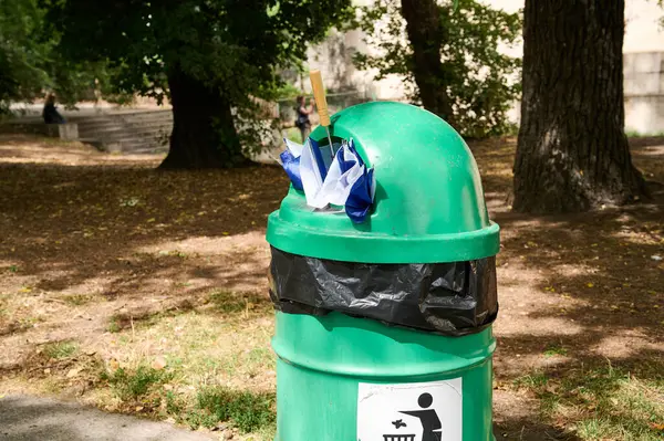 Siyah bir çantayla kaplı yeşil bir çöp tenekesi ağaçlarla çevrili bir parkta duruyor ve insanlar günlerinin tadını çıkarıyor..