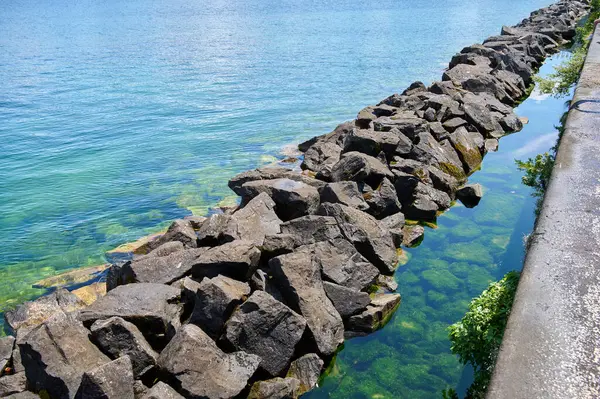 Büyük kayalar, güneşli bir günde temiz su ve yeşil bitkilerle çevrili kıyı boyunca bir bariyer oluşturur..
