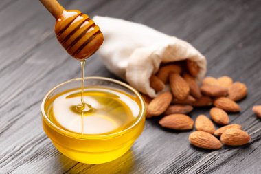 Honey is poured from a dipper into a glass bowl and almonds poured out of a bag on a wooden background. Still life of honey and scattered nuts on an aged wooden background.