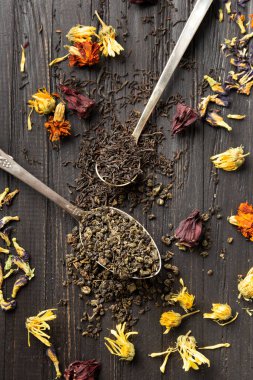 Composition of dry black and green tea in vintage spoons and dry hibiscus, calendula flowers on a dark wooden background top view close-up. Different types of dry tea on a dark background.