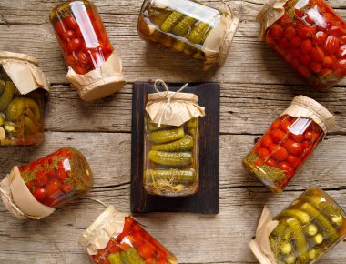 Canned cucumbers and tomato in glass jars on an old wooden background top view. Canned cherry tomatoes and carnishons on a wooden background.
