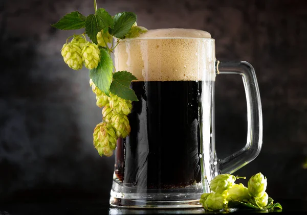 dark beer in a mug and green hop on wooden table on brown background