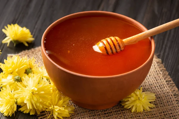 Honey in a clay bowl, dipper and yellow flowers on a textile background. A stick for honey lies in a clay bowl with honey close-up. Composition of honey in a plate and flowers on a dark wooden background.