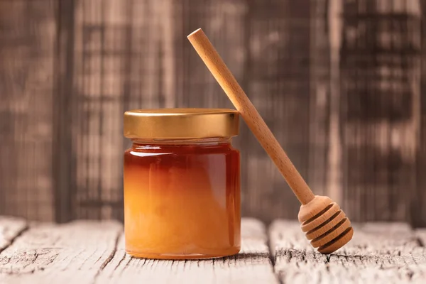 Closed jar with buckwheat honey and wooden dipper on a wooden background close-up. Composition of a jar of honey and a honey stick leaning against it.
