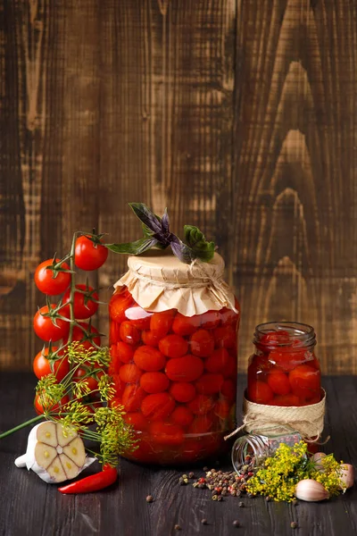 Pickled cherry tomatoes with garlic, dill and spices. Homemade food. Canned tomatoes in closed and open jars with a branch of fresh cherry tomatoes on a wooden background. Pickled vegetables.