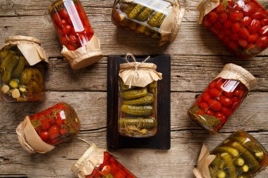 Canned cucumbers and tomato in glass jars on an old wooden background top view. Canned cherry tomatoes and carnishons in jars with kraft lids on a wooden background.
