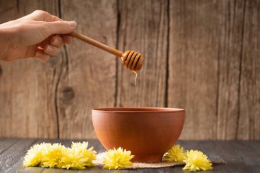 A hand with a dipper pouring honey into a clay bowl. Honey is poured from a dipper into a bowl on a wooden background close-up. Composition of pouring honey, honey stick and yellow flowers on a wooden background.