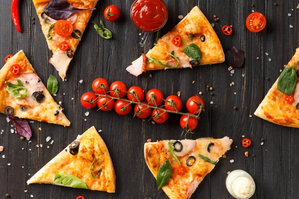 Pizza slices with salami, jerky, olives, cheese and herbs are laid out on a dark wooden background, a branch of fresh cherry tomatoes, sauces and seasonings for pizza top view. Italian food. Pizza on a black table.