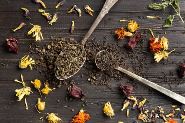 Composition of dry black and green tea in vintage spoons and dry hibiscus, calendula flowers on a dark wooden background top view close-up. Different types of dry tea on a dark background.