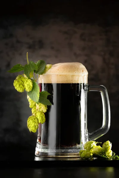 dark beer in a mug and green hop on wooden table on brown background. Oktoberfest. Beer Festival.