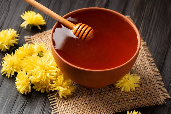 Honey in a clay bowl, dipper and yellow flowers on a textile and wooden background. A stick for honey lies in a clay bowl with honey close-up. Composition of honey in a plate and flowers on a dark background.