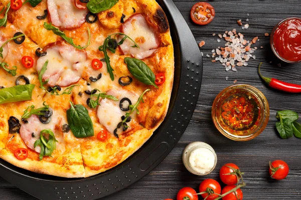 Pizza with salami, olives, cheese, herbs on a black dish, sauce, olive oil with spices and fresh vegetables on a dark wooden background. Homemade pizza with meat and herbs. Traditional Italian food.