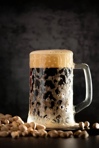 A glass of dark beer with foam on a dark background and a handful of pistachio nuts. Pistachios and beer. Foam pours over a glass of beer. Beer Festival. Octoberfest.