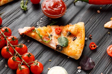 A slice of pizza with jerky, olives, cheese and herbs lies on a dark wooden background, a branch of fresh cherry tomatoes, pizza sauce. Italian food. Pizza on a black table.