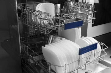 Built-in dishwasher in the kitchen with the door open and clean dishes inside. Clean white plates, saucepan, glasses are stacked inside the dishwasher. Household appliances for the kitchen.