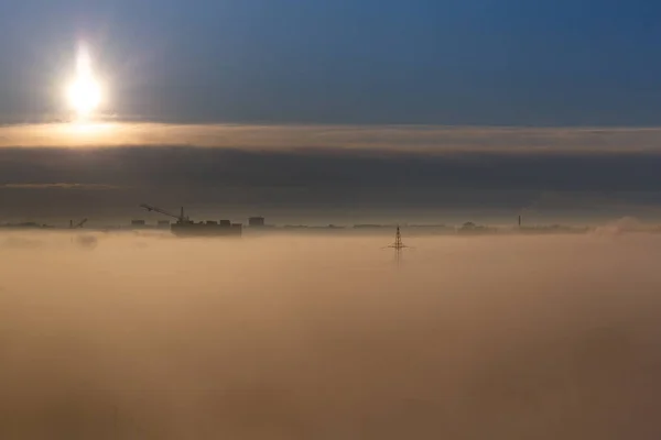 Sunrise in the city, foggy morning, silhouettes of multi-storey buildings, construction cranes and high-voltage poles in the fog. City landscape at dawn. Thick low fog and bright rising sun.