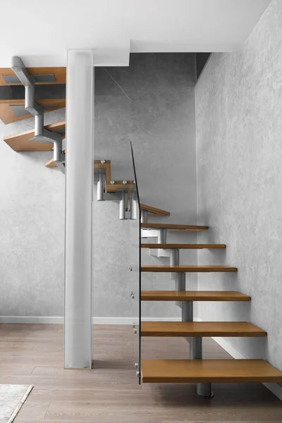 A staircase with wooden steps and an elegant glass fence, a column of matte glass in the modern interior of the apartment. Stairs design in a two -level apartment.