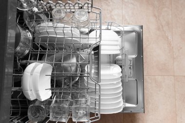 Built-in dishwasher in the kitchen with the door open and clean dishes inside. Clean white plates, saucepan, glasses are stacked inside the dishwasher. Household appliances for the kitchen.