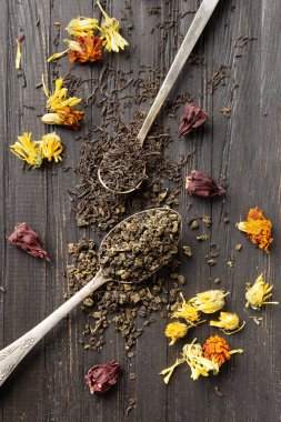 Antika kaşıklardaki kuru siyah ve yeşil çay kompozisyonu, koyu ahşap arka planda yakın plan görüntüsünde kuru çiçek çayı. Koyu arkaplanda farklı türde kuru çay..