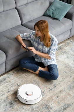 Bir kız oturma odasında yerde oturuyor ve akıllı telefon kullanarak robot elektrik süpürgesini kontrol ediyor. Akıllı Ev. Akıllı ev aletleri, ev işleri ve teknoloji kavramı..