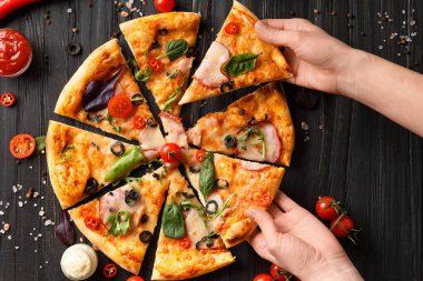 Eller, koyu ahşap arka planda kesilmiş pizzadan dilimler alır. Salamlı pizza, kurutulmuş et, sos, baharat, siyah arka planda taze vişneli domates. Kadınların elleri pizzayı paylaşır..