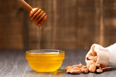 Honey is poured from a dipper into a glass bowl and almonds poured out of a bag on a wooden background. Still life of honey and scattered nuts on an aged wooden background.