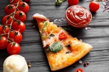 Kurutulmuş, zeytinli, peynirli ve otlu pizza dilimleri koyu ahşap arka planda, taze vişneli domates, sos ve pizza için baharatlardan oluşan bir dal üzerine serilir. İtalyan yemeği..