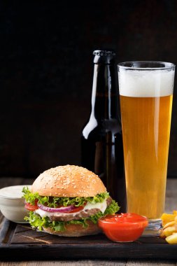 Sulu sığır etli burger, patates kızartması, iki sos, koyu renkli bir şişede bira ve siyah bir tahtada bira kupası ve ahşap arka plan. Bir barda Amerikan fast food, yiyecek ve içeceği..