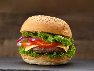 İştah açıcı burger, sulu sığır pirzolası, peynir, domates, marul, siyah ahşap zemin üzerinde siyah bir taş tabakta soğan. Fast food, ev yapımı hamburger..
