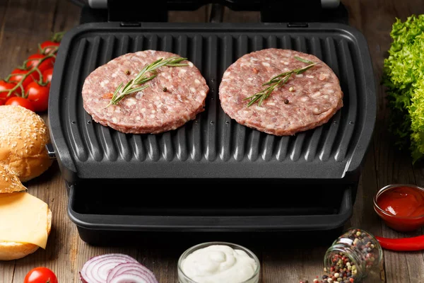 素朴な木製の背景にスマートな電気グリルでバーガーのための牛肉のパテが揚げられています 自家製ハンバーガーを作る ハンバーガーの具材 — ストック写真