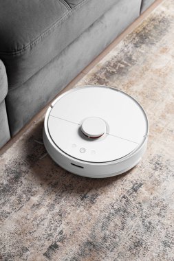 Kanepenin yanındaki oturma odasındaki halıda beyaz robot süpürgesi var. Daireyi modern bir elektrikli süpürgeyle temizliyorum..