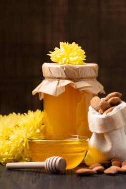 Honey in a closed glass jar and a bowl, sticks for honey and scattered almonds on a wooden background. Composition of honey jar, dipper, nuts in a bag close-up.