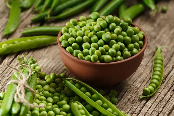 Fresh organic green peas in closed and open pods, scattered pea seeds, shelled green peas in a clay bowl on an aged wooden background. vegetable protein.