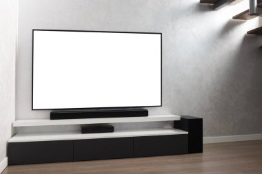 Oturma odasının içinde duvarında beyaz ekran televizyon, hifi ekipmanları, ses çubuğu, gri dolap olan bir yer. TV ve müzik sistemi iç mekanda.