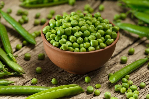 Fresh organic green peas in closed and open pods, scattered pea seeds, shelled green peas in a clay bowl on an aged wooden background. vegetable protein.