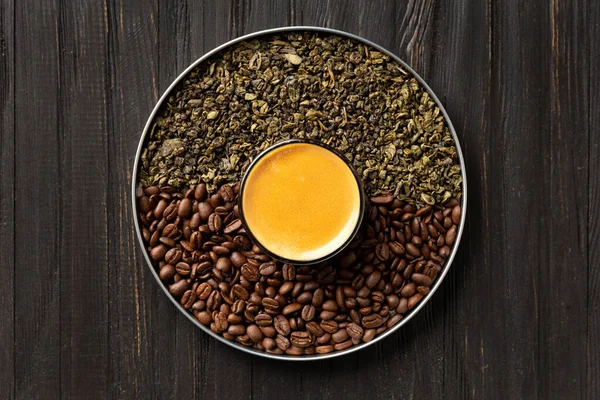 Composition of dry green tea leaves, roasted coffee beans in a round box and coffee in a cup on a dark wooden background top view.