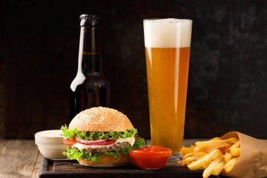 Burger, beer in a bottle and in a glass mug, french fries, sauce on a dark background, fast food concept.