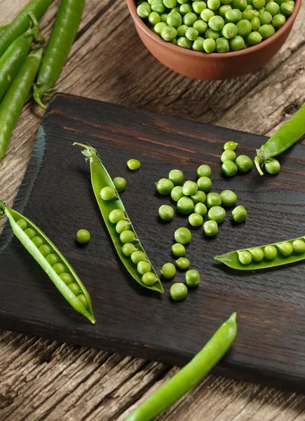 Fresh organic green peas in closed and open pods, scattered pea seeds, peeled green peas in a clay bowl on a dark wooden board and aged wooden background. vegetable protein.