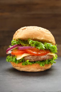 İştah açıcı burger, sulu sığır pirzolası, peynir, domates, marul, siyah ahşap zemin üzerinde siyah bir taş tabakta soğan. Fast food, ev yapımı hamburger..