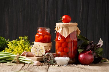 Kapalı ve açık kavanozlarda konserve kiraz domatesleri, ahşap arka planda salamura için baharatlar ve otlar..