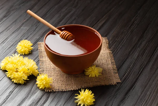Honey in a clay bowl, dipper and yellow flowers on a textile and a dark wooden background. A stick for honey lies in a ceramic bowl with honey close-up.