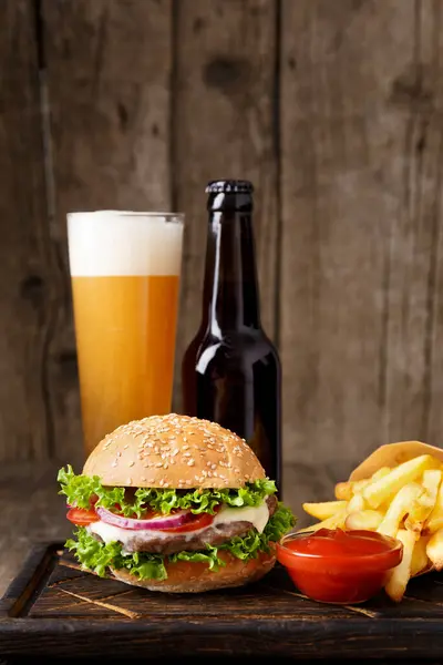 Burger, beer in a bottle and in a glass mug, french fries, sauce on a wooden background, fast food concept.