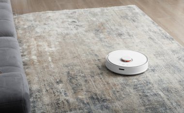 Kanepenin yanındaki oturma odasındaki halıda beyaz robot süpürgesi var. Daireyi modern bir elektrikli süpürgeyle temizliyorum..