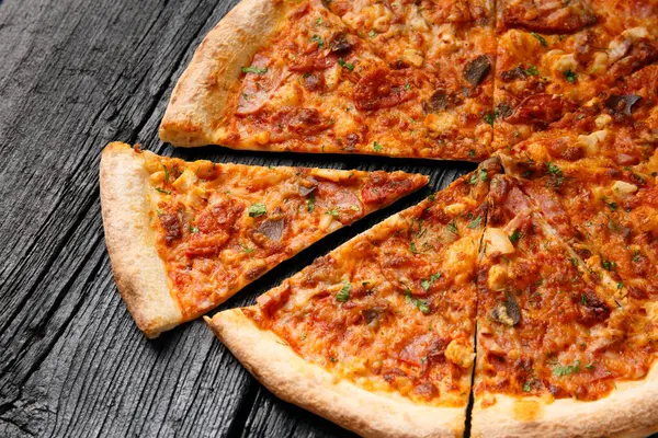 Pizza with different types of meat, cut into slices, one slice is separated on a black wooden background