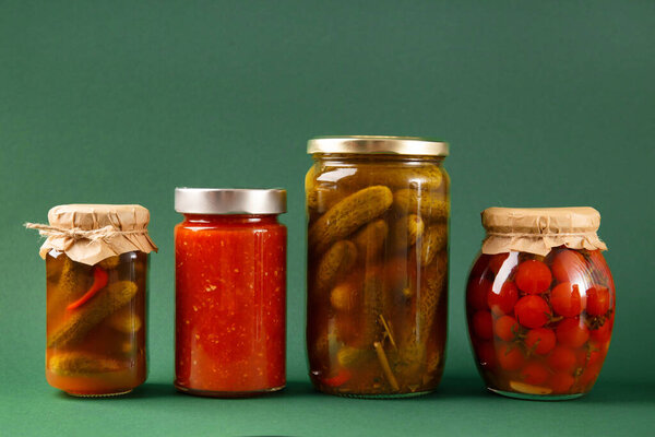 Canned vegetables in closed glass jars on a green background. Pickled cucumbers, tomatoes, chili sauce, adjika. Spicy snack, home canned food.
