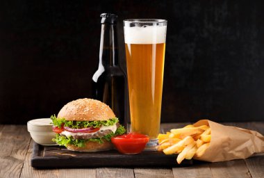 Burger, beer in a bottle and in a glass mug, french fries, sauce on a dark background, fast food concept.