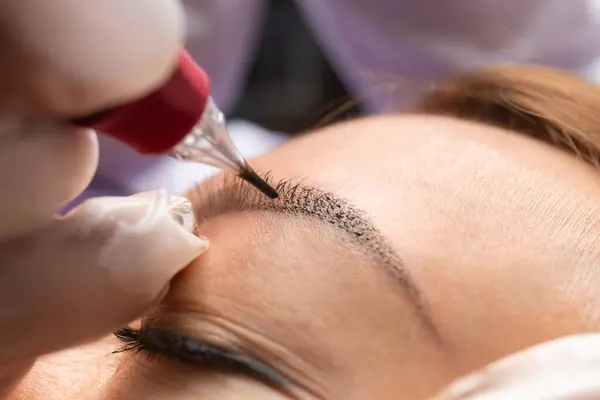 Beauty master performs permanent eyebrow makeup in a beauty salon. Hands of a cosmetologist doing microblading of eyebrows. Tattoo on the face.