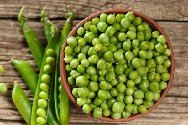 Peeled green peas in a clay bowl, sweet organic green peas in closed and open pods and scattered grains on an aged wooden background, top view. vegetable protein.