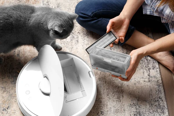 The container of the vacuum cleaner robot is in female hands, the dust collector of the robot vacuum cleaner is full of wool, the cat sits nearby and looks at the container.